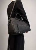 A dark gray textured leather shoulder bag with top handles and a front zip pocket, shown worn crossbody over a black dress against a neutral gray background.