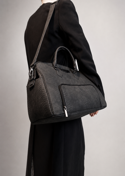 A dark gray textured leather shoulder bag with top handles and a front zip pocket, shown worn crossbody over a black dress against a neutral gray background.