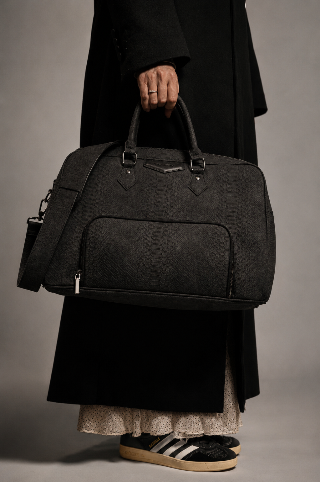A person in a long black coat carrying the Lacey Louise Liberty bag, a black textured leather duffel with top handles and a front zip pocket.